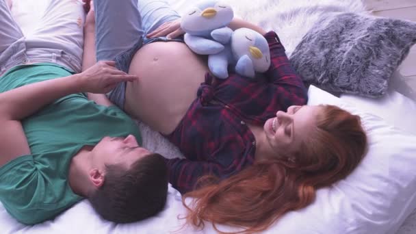 Kobieta w ciąży z długimi włosami i jej mąż, leżąc na łóżku. Mężczyzna naśladuje czynności palcami — Wideo stockowe