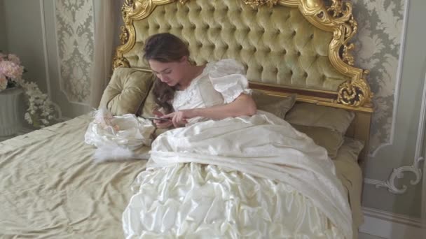 Jovem mulher de vestido de baile deitada na cama decorada a ouro e textos por telefone celular. Menina usa gadget — Vídeo de Stock
