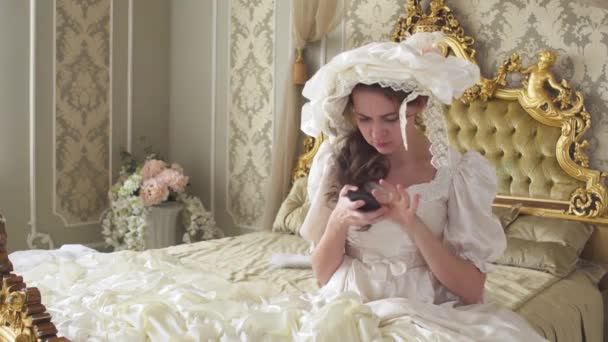 Young woman in ball gown sits on gold decorated bed and texts on cell phone. Princess uses gadget — Stock Video