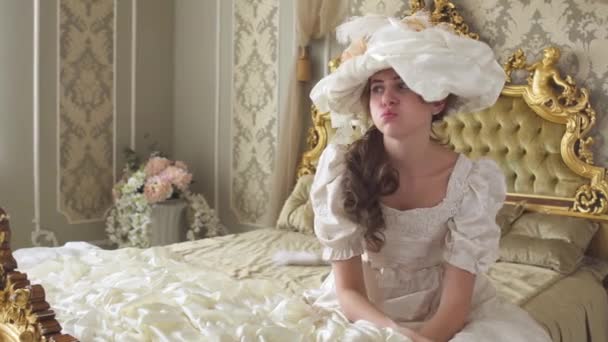 Jovem mulher entediada em vestido de baile e chapéu sentado na enorme cama dourada e jogar chapéu no chão — Vídeo de Stock