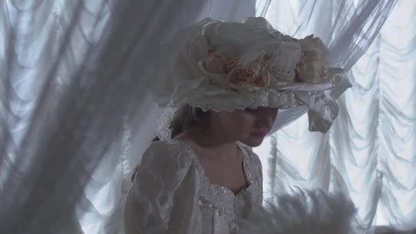 Jolie jeune femme en robe de bal vintage blanche et chapeau blanc debout sur le fond de rideaux transparents et agitant ventilateur parlant sur un téléphone portable — Video