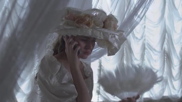Портрет досить молодої усміхненої жінки в білій вінтажній кульковій сукні та білому капелюсі, що стоїть на тлі прозорих штор та махає вентилятором, що розмовляє на мобільному телефоні — стокове відео