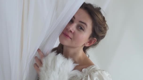 Portrait young shy playful woman in vintage ball costume lookng through white veil holding feather fan in hand on white background half face real people series. — Stock Video