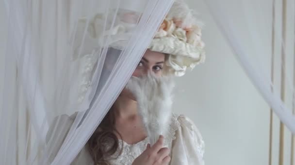 年轻美丽的笑脸女人调情附近的薄纱挥舞着白色的羽毛风扇 女孩穿白色礼服和白色帽子与鲜花 — 图库视频影像