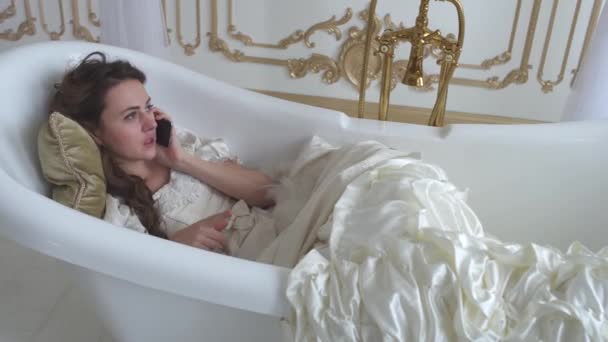 Красивая молодая женщина лежит в ванне на мягкой подушке и разговаривает по мобильному телефону с вентилятором перьев в руках — стоковое видео