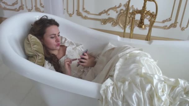 Ung kvinna i balklänning ligga i badet med kudde och texter på mobiltelefon. Flicka använder gadget — Stockvideo