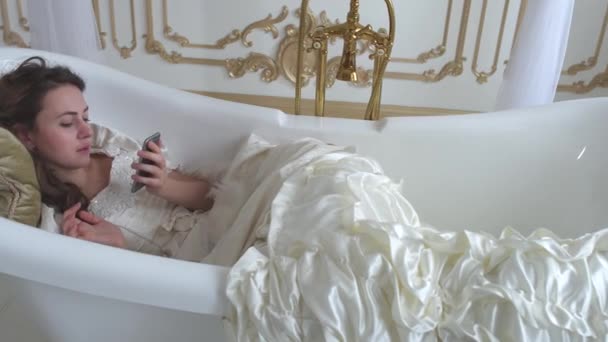 Милая молодая женщина лежит в ванне на мягкой подушке и разговаривает по мобильному телефону с перьевым вентилятором в руках — стоковое видео