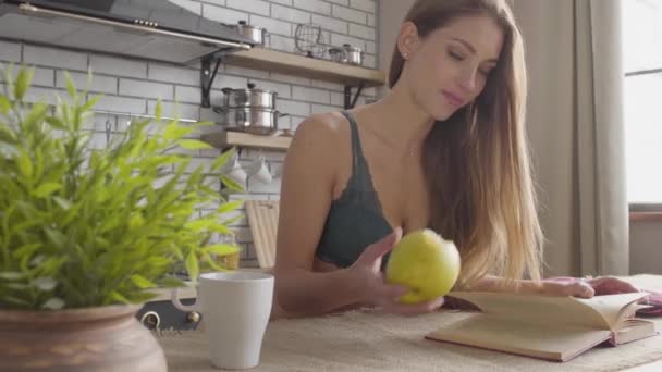 Retrato mulher bonita em um livro de leitura de sutiã e comer maçã verde na cozinha. Estilo de vida saudável da jovem. Lazer de jovem bela menina solitária . — Vídeo de Stock