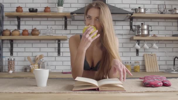 Retrato jovem sensual mulher em lingerie ler livro e comer maçã verde na cozinha. Estilo de vida saudável da jovem. Lazer de jovem bela menina solitária . — Vídeo de Stock