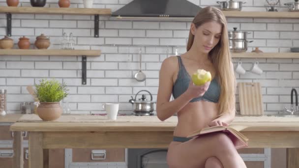 Красива жінка в нижній білизні сидить на кухні читаючи книгу і їдячи зелене яблуко. Здоровий спосіб життя красивої леді. Дозвілля молодої красивої самотньої дівчини . — стокове відео
