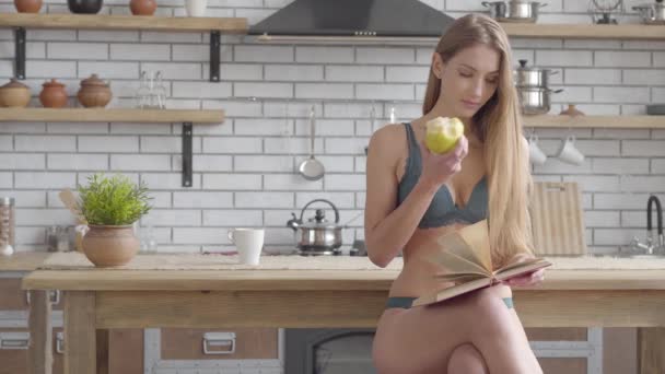 Retrato bastante joven mujer en lencería sentado en el libro de lectura de la cocina y comer manzana verde. Un estilo de vida saludable de hermosa dama. Ocio de joven hermosa chica solitaria . — Vídeos de Stock