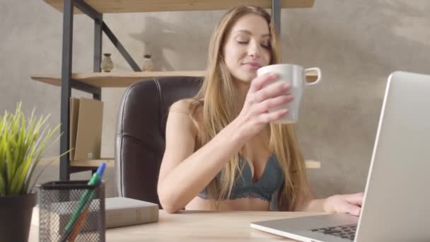Portrét radost radostné hezká mladá žena na prádlo černé kožené židli u stolu psaní na svém laptopu, pít kávu nebo čaj z hrnečku bílého v moderní kanceláři.