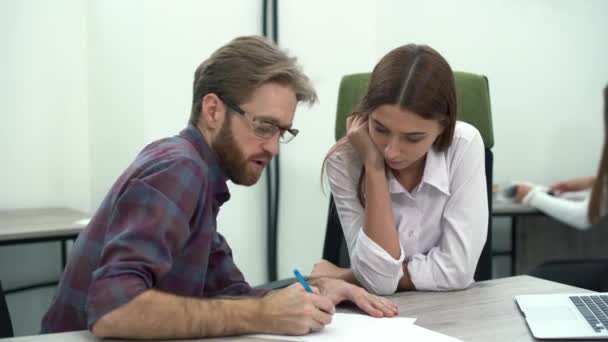 Gerente barbudo con gafas escribiendo en un plan de desarrollo de papel mientras está sentado en la mesa con su empleado. Reunión creativa del equipo de negocios en la oficina moderna . — Vídeo de stock