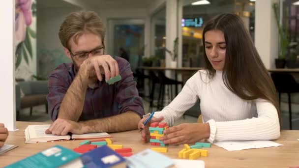 Attractive bearded man with glasses and a cute girlfriend with long dark hair are building a tower of multi-colored wooden blocks while sitting at the table. Friends play an interesting strategy game. — Stock Video