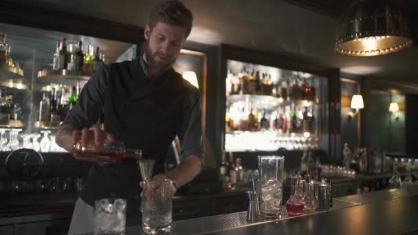Высокий бородатый бармен наливает ром в металлическую банку, затем в стекло. Бармен делает коктейль в современном баре с большим количеством бутылок на полках — стоковое видео