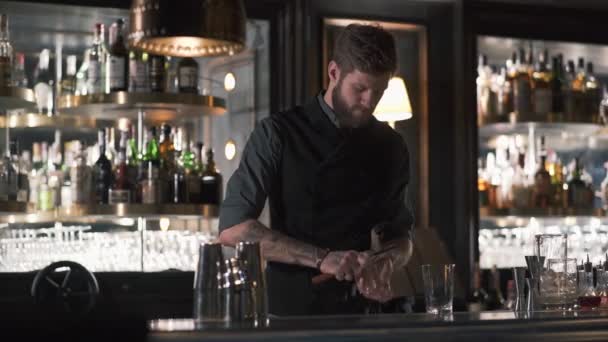 Höga skäggiga bartender krossning stor bit av is med kniv står i baren motverka. Bartendern att göra cocktail i modern bar med många flaskor på hyllor — Stockvideo