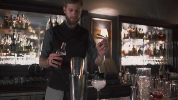 酒保把酒精倒在烧杯里, 然后倒在冰杯里, 展示了他的技能。巴曼在站在柜台附近的现代酒吧里做鸡尾酒。为客户准备酒精饮料的人 — 图库视频影像