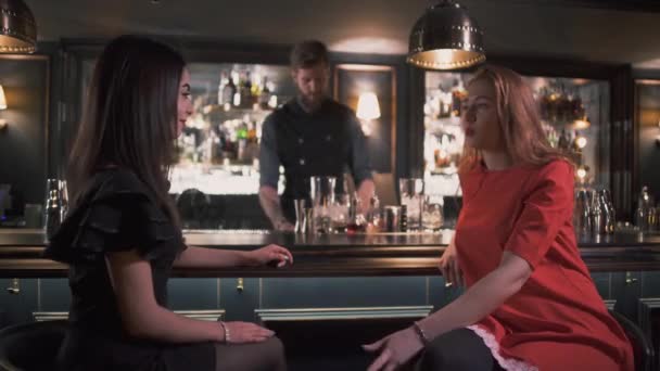 Deux copines assises près du comptoir du bar ont une conversation intéressante en gros plan. Barman en arrière-plan faire un cocktail pour le client, souriant. Loisirs de belles filles. dames solitaires avoir du plaisir — Video