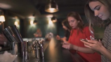 Üç meşgul genç kızlar oturup tarafından bar kokteyller için bekleyen ve onların smatphones mesajlar yazarak ve gülümseyen.