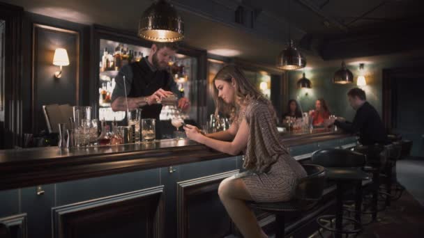 Бармен-хипстер сочетает ингредиенты и делает коктейли в баре, в то время как молодая расстроенная женщина сидит возле барной стойки и печатает смс — стоковое видео
