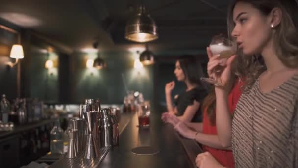 Tres hermosas chicas jóvenes en vestidos de noche sentados en el mostrador del bar y bebidas cóctel — Vídeo de stock