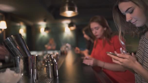 Tre upptagen unga flickor sitta vid baren väntar cocktails och skriva meddelanden i sin smatphones och leende. — Stockvideo
