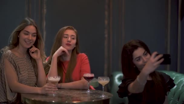 Три симпатичные девушки сидят в кафе или ресторане красиво позируют и делают фото селфи для социальных сетей — стоковое видео