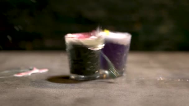 Dos vasos con bebidas alcohólicas multicolores chocan sobre la mesa y se rompen en fragmentos. Movimiento lento . — Vídeo de stock