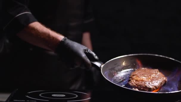 Male hand in black rubber gloves holding a frying pan with a grilling steak. The cook turns the pan on the fire in different directions. Slow motion. — Stock Video