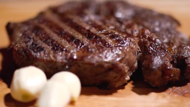 Heerlijke gegrilde steak liggend op de snijplank met knoflook close-up. Verse rozemarijn vallen op het vlees — Stockvideo