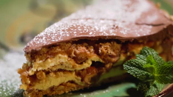 Смачний апетитний шматок торта з горіхами та шоколадом, посипаний солодким порошком, що лежить на тарілці поруч з листя м'яти — стокове відео