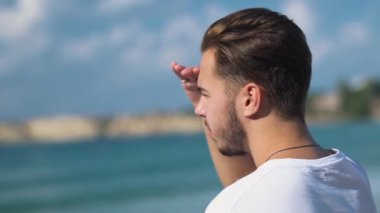 Su almak üzerinde Güneş'ten el gözleriyle kapsayan deniz maliyet üzerinde duran yakışıklı delikanlı. Beyaz t-shirt denizde dinlenme sakallı adam. Yalnız adamın boş zaman
