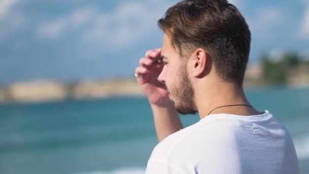 Retrato de cerca del perfil del joven guapo alisándose el pelo y centrado en mirar el mar sobre el fondo de la playa — Vídeos de Stock