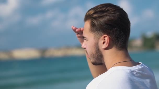 ハンサムな若い男が水の上を探して、太陽から手で彼の目を覆っている海コストの上に立って。海で白い t シャツで髭の男。孤独な男のレジャー — ストック動画