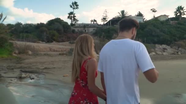 Прекрасная пара прогуливается вдоль берега моря, держа руки близко. Высокий мужчина в белой футболке и женщина в красном платье отдыхают вместе. Камера движется по кругу — стоковое видео