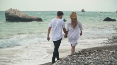 Arkadan görünüşü güzel genç çift sahilde elele ve taşlar üzerinde yürüme. Sıçramasına dalgalar deniz. Şaşırtıcı görünümü. Ağır çekim.