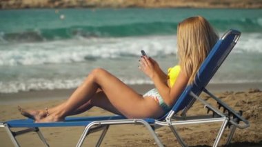 Plajda şezlong, arka plan denizde yatan güneşlenme sarışın bir kadın. Kız manifatura onun cep telefonu. Eğlence genç kadın