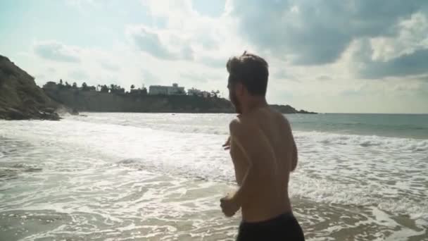 Hombre deportivo corriendo con el torso desnudo y pantalones negros corriendo en la pintoresca hermosa playa de mar en el día soleado. Movimiento lento — Vídeos de Stock