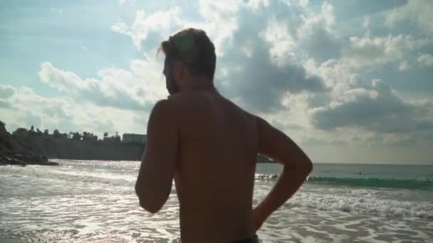 Mladý muž běží podél pobřeží ráno. Muž otočí na cestách a dívá se do kamery. Běhání v blízkosti moře v pomalém pohybu. Zdravý životní styl. Léto na pláži — Stock video