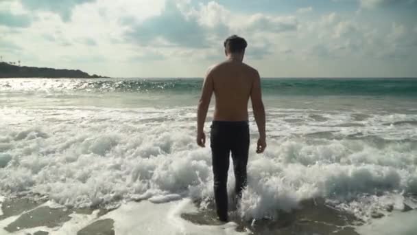 ジーンズの若い男は スローモーションでカメラに背中を浅い海の水を入る 泡波は 海岸にロールバックします 夏のビーチで — ストック動画