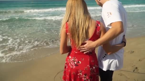 Молодая пара стоит на пляже, любуясь красивым морским пейзажем, мужчина и женщина обнимаются. Человек нарисовал сердце и почерк любви на посылке с палкой. Стреляли сзади. Камера опускается. — стоковое видео