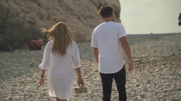 Pareja joven caminando por la playa con piedras sin zapatos, y el pelo y la ropa ligera se balancean en el viento. Movimiento lento — Vídeo de stock