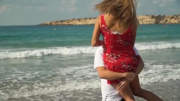 Bebaarde man verhoogd het meisje in jurk in zijn armen en spinnen rond met haar op het strand. Gelukkige paar tijd samen in de buurt van de Oceaan. Vrije tijd in de zomer — Stockvideo