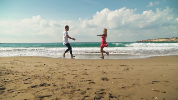 年轻的一男一女在海滩上向对方跑去。男人把那个抱着衣服的女孩抱起来, 和她一起旋转。可爱的恋人快乐在一起。夏天的休闲。慢动作. — 图库视频影像
