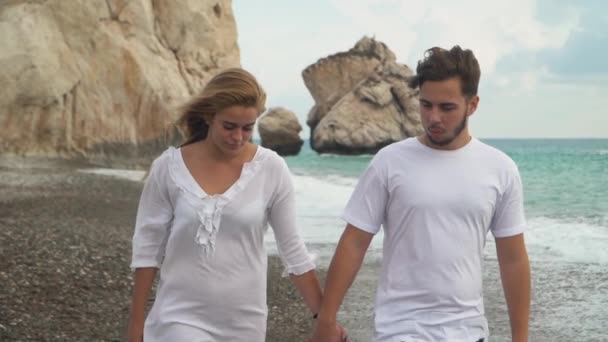 Ein junges Paar in weißen Kleidern geht entlang der felsigen Küste in der Nähe des Meeres oder Ozeans und kommuniziert Händchen haltend miteinander. Zeitlupe. — Stockvideo