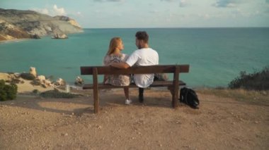 Genç çift sarılma temiz su ile sonsuz denizin muhteşem bir ısmarlayarak bir uçurumun üzerinde bir bankta oturuyor