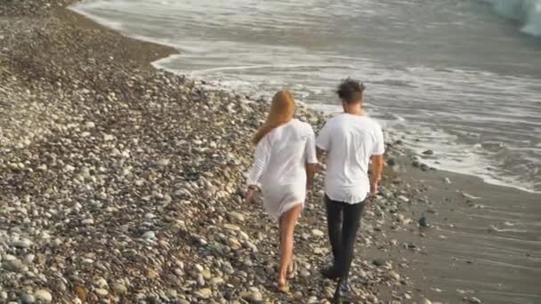 白いドレスで幸せなカップルは、手を繋いでの海岸に沿って歩いてください。若い家族のレジャー。ドローンは、背面から撮影します。トップ ビュー — ストック動画