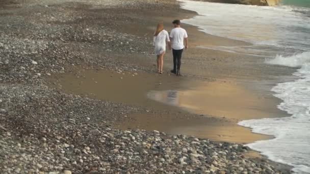 Pareja feliz vestida de blanco camina a lo largo de la costa rocosa tomados de la mano lejos. Ocio de familia joven. Disparando por la espalda. Vista superior — Vídeos de Stock