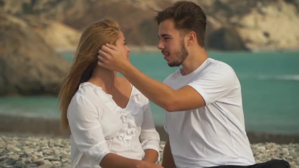 Unga paret sitter varma stenar på stranden, de glatt kommunicera, och killen avlägsnar skonsamt flicka s håret bakom örat — Stockvideo