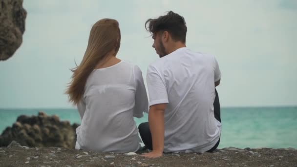 ΠΙΣΩ ΟΨΗ νεαρό όμορφο ζευγάρι κάθεται σε ζεστό βότσαλα στην παραλία, που ευτυχώς επικοινωνούν και να περνούν χρόνο μαζί. — Αρχείο Βίντεο